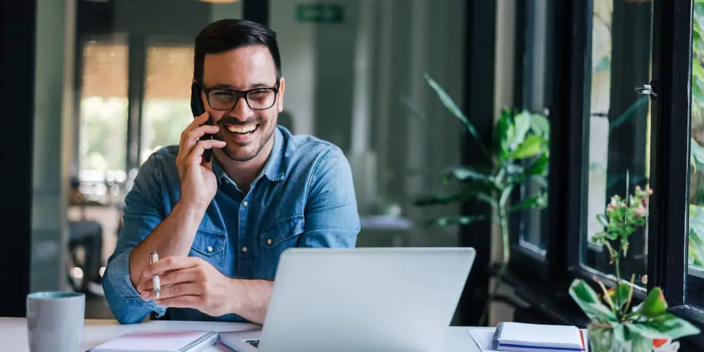 un empresario sonriente utiliza las habilidades interpersonales mientras habla por teléfono con su portátil delante